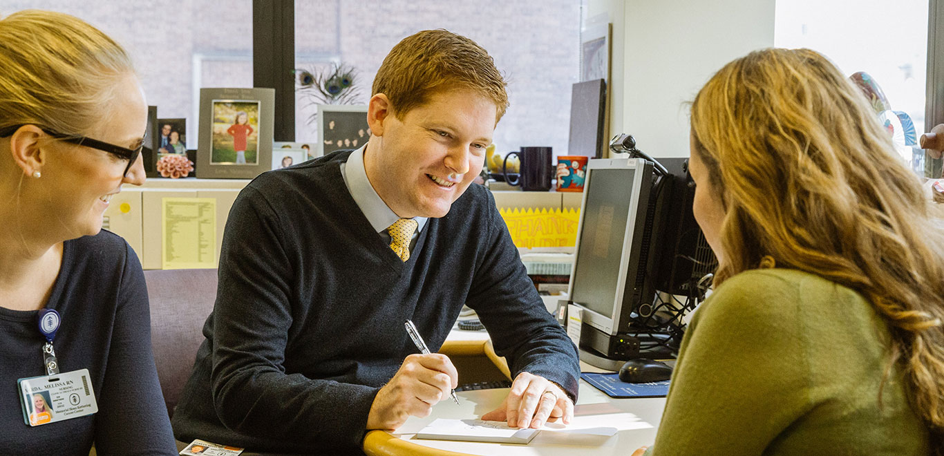 Man in jumper vest sat at a desk chatting to another person and writing things down